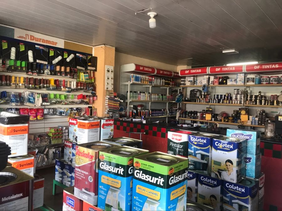 Interior da DF Tintas: pilhas de latas de tinta, prateleiras com a faixa vermelha DF Tintas e parede de rolos e pincéis