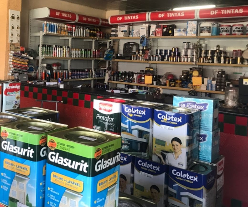 Prateleiras com a faixa vermelha DF Tintas e latas de Glasurit, Colatex, Resicolor e Iquine Pintalar na frente do balcão xadrez vermelho e verde